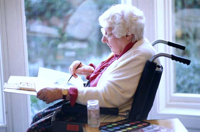 elderly woman in a wheelchair painting beside a window. 