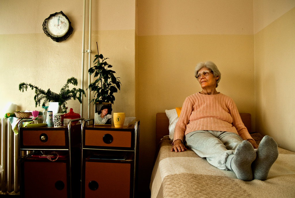 Elderly woman in an orange sweater sitting on her bed in a nursing home