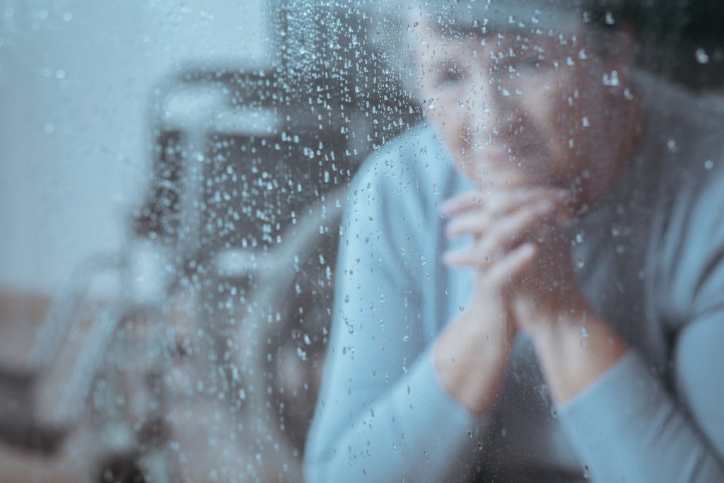Depressed elderly woman looking through a rainy window with a wheelchair behind her