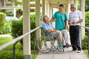 an older man in a wheel chair with a nurse and another older man standing with them having a conversation outside. 