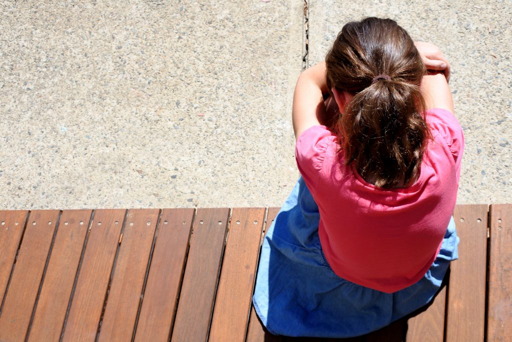 Overhead view of sad young girl (age 9-10) covering her face and crying in school yard.