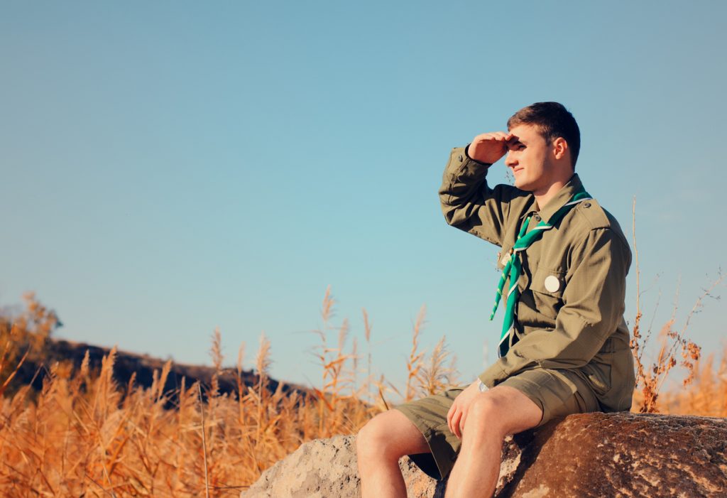 Young Boy Scout in Uniform Watching Over a Field on A Sunny Day While Sitting on a Boulder