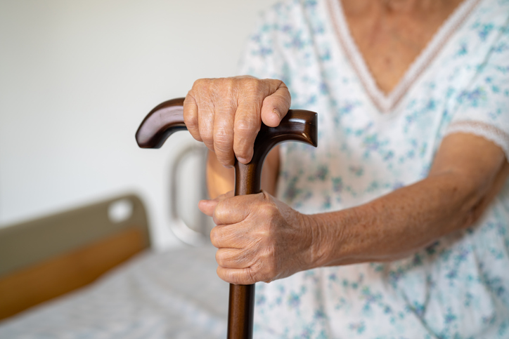 Woman with cane in nursing home sitting on bed.